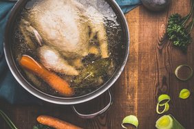 Chicken soup cooking, pot with chicken broth and ladle on dark rustic wooden background with vegetables ingredients, top view. Healthy food or Diet nutrition concept